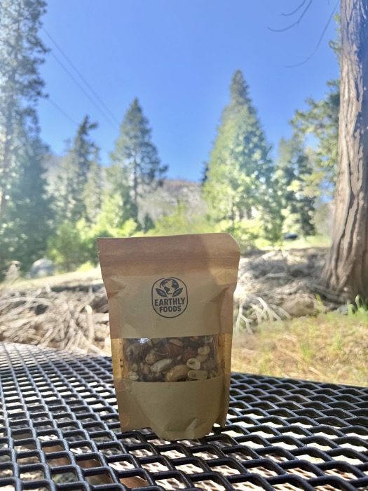 A bag of Earthly Foods trail mix on a picnic table in a pine forest, with a fallen log in the background.