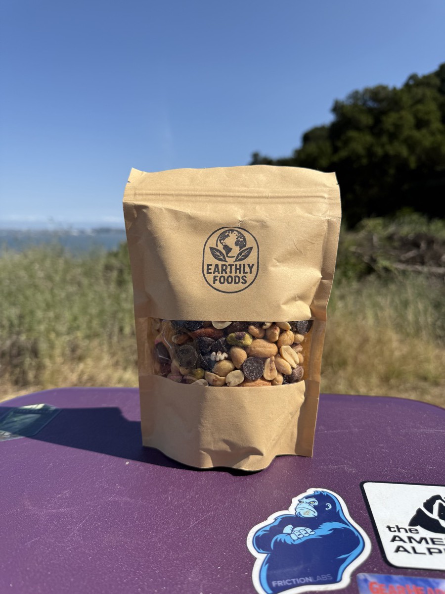 A bag of Earthly Foods trail mix on a picnic table with a bay and blue sky in the background.