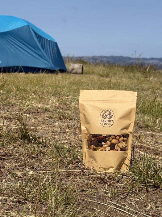 A bag of Earthly Foods trail mix in a grassy field with a blue camping tent in the background.
