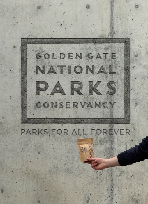 A hand holding a bag of Earthly Foods trail mix in front of a Golden Gate National Parks Conservancy sign.